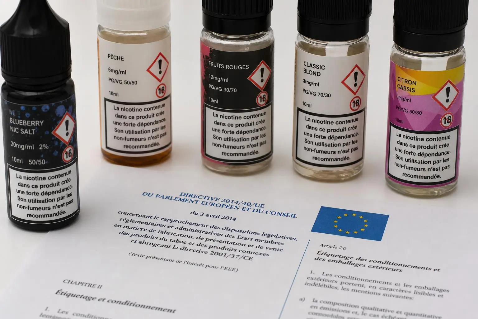 Cinq flacons de e-liquide avec étiquettes d’avertissement et un document européen à côté, axes de vapotage et risques. nicotine cigarette électronique danger évoqué.